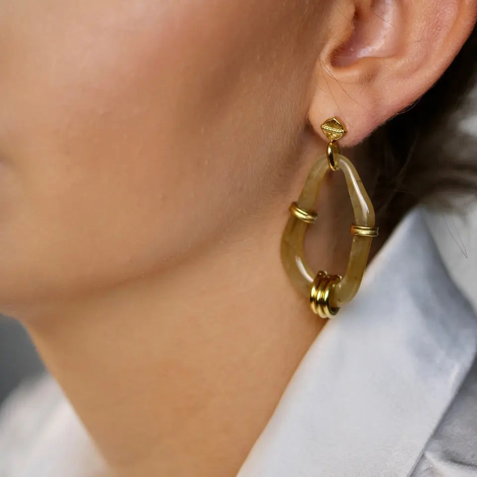 Boucles d’oreilles pendantes en résine ovaloïde beige ornées d’anneaux en acier inoxydable doré portées aux oreilles