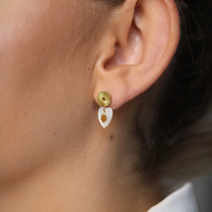 Boucles d’oreilles puces nacre en acier inoxydable doré composées d'une pièce pendante en nacre en forme de cœur orné d’un soleil portées aux oreilles