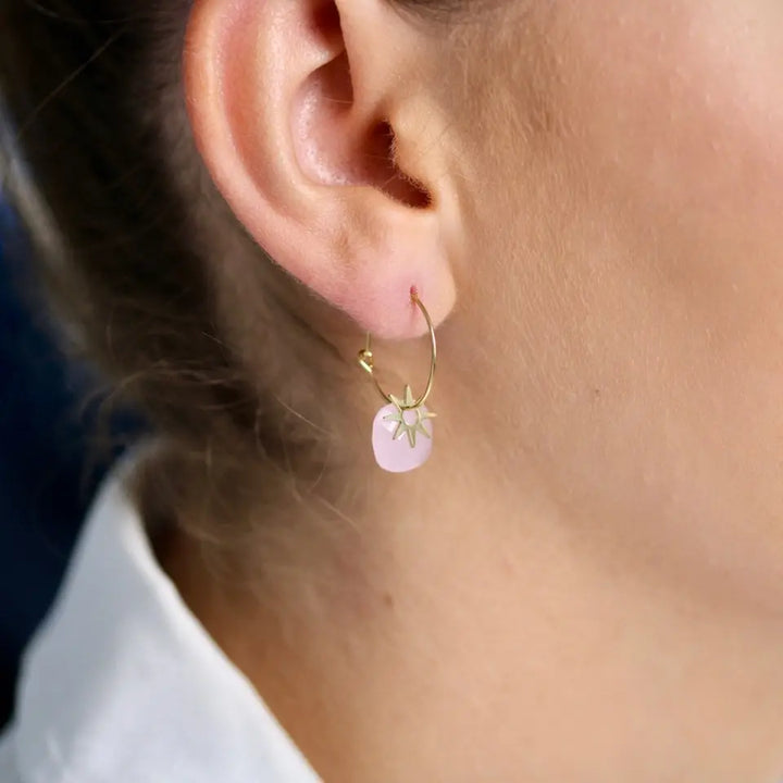 Boucles d’oreilles mini créoles avec pendentifs composées d’une pierre naturelle rose et d’un motif en forme de soleil doré en acier inoxydable portées aux oreilles